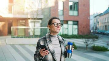 retrato de un joven caucásico mujer de negocios con lentes y un Saco camina mediante el negocio distrito, Bebiendo café y utilizando teléfono inteligente video