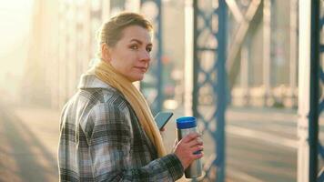 portrait de une Jeune caucasien femme d'affaires dans une manteau, en marchant à travers le pont sur une glacial ensoleillé matin, en buvant café et en utilisant téléphone intelligent. communication, travail jour, occupé la vie concept video