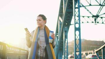 portret van een jong Kaukasisch zakenvrouw in een jas, wandelen aan de overkant de brug Aan een ijzig zonnig ochtend, drinken koffie en gebruik makend van smartphone. communicatie, werk dag, bezig leven concept video