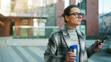 retrato de un joven caucásico mujer de negocios con lentes y un Saco camina mediante el negocio distrito, Bebiendo café y utilizando teléfono inteligente video