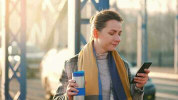 porträtt av en ung caucasian affärskvinna i en täcka, gående tvärs över de bro på en frostig solig morgon, dricka kaffe och använder sig av smartphone. kommunikation, arbete dag, upptagen liv begrepp video