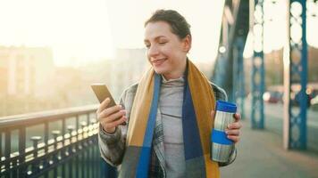 retrato de un joven caucásico mujer de negocios en un abrigo, caminando a través de el puente en un escarchado soleado mañana, Bebiendo café y utilizando teléfono inteligente comunicación, trabajo día, ocupado vida concepto video