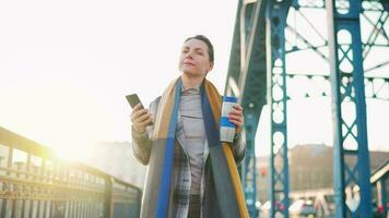 portret van een jong Kaukasisch zakenvrouw in een jas, wandelen aan de overkant de brug Aan een ijzig zonnig ochtend, drinken koffie en gebruik makend van smartphone. communicatie, werk dag, bezig leven concept video