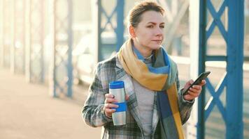 portret van een jong Kaukasisch zakenvrouw in een jas, wandelen aan de overkant de brug Aan een ijzig zonnig ochtend, drinken koffie en gebruik makend van smartphone. communicatie, werk dag, bezig leven concept video