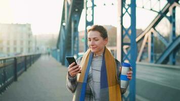 portret van een jong Kaukasisch zakenvrouw in een jas, wandelen aan de overkant de brug Aan een ijzig zonnig ochtend, drinken koffie en gebruik makend van smartphone. communicatie, werk dag, bezig leven concept video