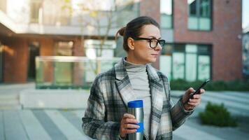 Porträt von ein jung kaukasisch Geschäftsfrau mit Brille und ein Mantel Spaziergänge durch das Geschäft Bezirk, Trinken Kaffee und mit Smartphone video