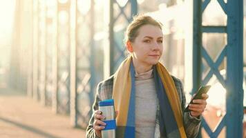 portret van een jong Kaukasisch zakenvrouw in een jas, wandelen aan de overkant de brug Aan een ijzig zonnig ochtend, drinken koffie en gebruik makend van smartphone. communicatie, werk dag, bezig leven concept video