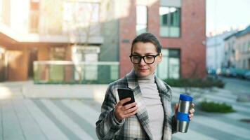 portret van een jong Kaukasisch zakenvrouw met bril en een jas wandelingen door de bedrijf wijk, drinken koffie en gebruik makend van smartphone video