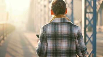 Portrait of a young caucasian businesswoman in a coat, walking across the bridge on a frosty sunny morning, drinking coffee and using smartphone. Communication, work day, busy life concept video