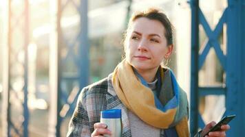 portrait de une Jeune caucasien femme d'affaires dans une manteau, en marchant à travers le pont sur une glacial ensoleillé matin, en buvant café et en utilisant téléphone intelligent. communication, travail jour, occupé la vie concept video