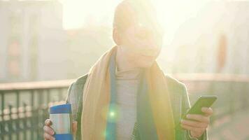 Portrait of a young caucasian businesswoman in a coat, walking across the bridge on a frosty sunny morning, drinking coffee and using smartphone. Communication, work day, busy life concept video