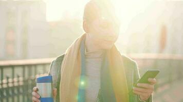 retrato de un joven caucásico mujer de negocios en un abrigo, caminando a través de el puente en un escarchado soleado mañana, Bebiendo café y utilizando teléfono inteligente comunicación, trabajo día, ocupado vida concepto video