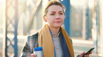 Portrait of a young caucasian businesswoman in a coat, walking across the bridge on a frosty sunny morning, drinking coffee and using smartphone. Communication, work day, busy life concept video