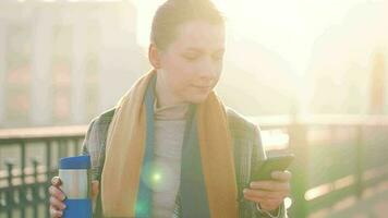 retrato de un joven caucásico mujer de negocios en un abrigo, caminando a través de el puente en un escarchado soleado mañana, Bebiendo café y utilizando teléfono inteligente comunicación, trabajo día, ocupado vida concepto video