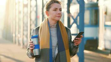 retrato de un joven caucásico mujer de negocios en un abrigo, caminando a través de el puente en un escarchado soleado mañana, Bebiendo café y utilizando teléfono inteligente comunicación, trabajo día, ocupado vida concepto video