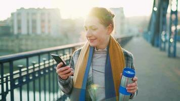 retrato de un joven caucásico mujer de negocios en un abrigo, caminando a través de el puente en un escarchado soleado mañana, Bebiendo café y utilizando teléfono inteligente comunicación, trabajo día, ocupado vida concepto video