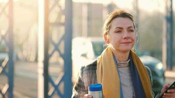 Portrait of a young caucasian businesswoman in a coat, walking across the bridge on a frosty sunny morning, drinking coffee and using smartphone. Communication, work day, busy life concept video