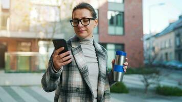 retrato de un joven caucásico mujer de negocios con lentes y un Saco camina mediante el negocio distrito, Bebiendo café y utilizando teléfono inteligente video