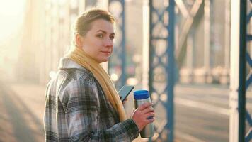 Porträt von ein jung kaukasisch Geschäftsfrau im ein Mantel, Gehen über das Brücke auf ein eisig sonnig Morgen, Trinken Kaffee und mit Smartphone. Kommunikation, Arbeit Tag, beschäftigt Leben Konzept video