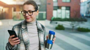 retrato de un joven caucásico mujer de negocios con lentes y un Saco camina mediante el negocio distrito, Bebiendo café y utilizando teléfono inteligente video