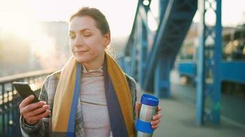 portret van een jong Kaukasisch zakenvrouw in een jas, wandelen aan de overkant de brug Aan een ijzig zonnig ochtend, drinken koffie en gebruik makend van smartphone. communicatie, werk dag, bezig leven concept video