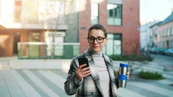 Portrait of a young caucasian businesswoman with glasses and a coat walks through the business district, drinking coffee and using smartphone video