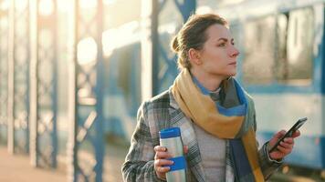 portret van een jong Kaukasisch zakenvrouw in een jas, wandelen aan de overkant de brug Aan een ijzig zonnig ochtend, drinken koffie en gebruik makend van smartphone. communicatie, werk dag, bezig leven concept video