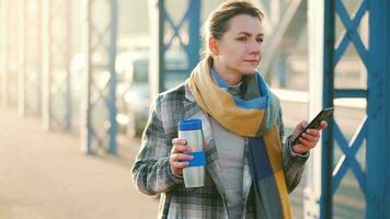 porträtt av en ung caucasian affärskvinna i en täcka, gående tvärs över de bro på en frostig solig morgon, dricka kaffe och använder sig av smartphone. kommunikation, arbete dag, upptagen liv begrepp video
