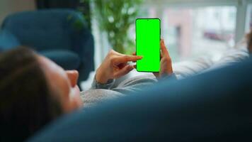 Woman at home using smartphone with green mock-up screen in vertical mode. Girl browsing Internet, watching content video