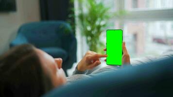 Woman at home using smartphone with green mock-up screen in vertical mode. Girl browsing Internet, watching content video