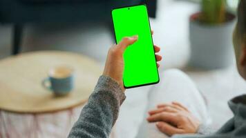 Woman at home using smartphone with green mock-up screen in vertical mode. Girl browsing Internet, watching content video