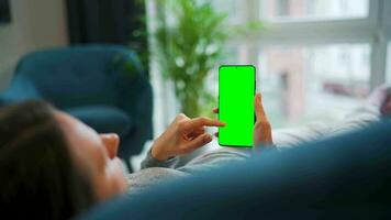 Woman at home using smartphone with green mock-up screen in vertical mode. Girl browsing Internet, watching content video