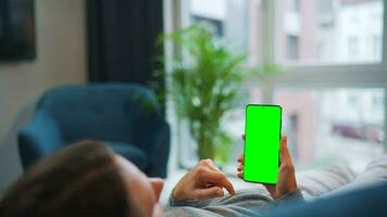 Woman at home using smartphone with green mock-up screen in vertical mode. Girl browsing Internet, watching content video