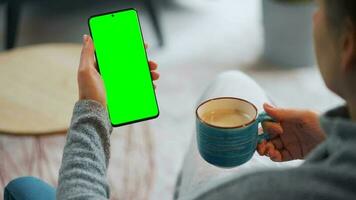 Woman at home using smartphone with green mock-up screen in vertical mode and drinking coffee video