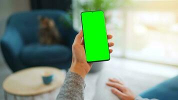 Woman at home using smartphone with green mock-up screen in vertical mode. Girl browsing Internet, watching content video