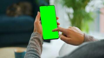 Woman at home using smartphone with green mock-up screen in vertical mode. Girl browsing Internet, watching content video