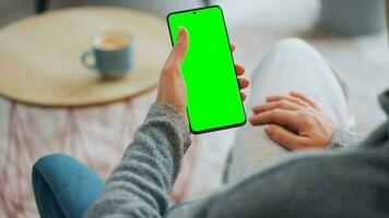 Woman at home using smartphone with green mock-up screen in vertical mode. Girl browsing Internet, watching content video