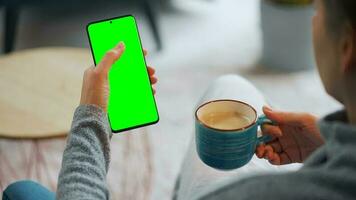 Woman at home using smartphone with green mock-up screen in vertical mode and drinking coffee video