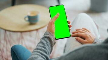 Woman at home using smartphone with green mock-up screen in vertical mode. Girl browsing Internet, watching content video