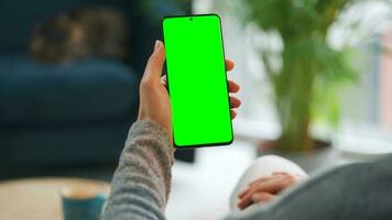 Woman at home using smartphone with green mock-up screen in vertical mode. Girl browsing Internet, watching content video