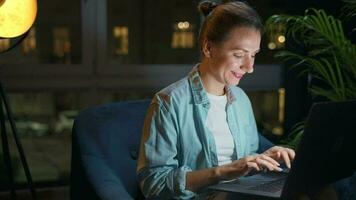 sorridente mulher é sentado dentro a poltrona e trabalhando em uma computador portátil ou conversando com alguém às noite. conceito do controlo remoto trabalhos video