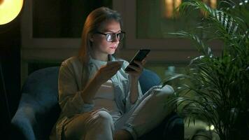 Woman is sitting in the armchair and makes an online purchase using a credit card and smartphone at night video