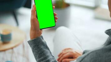 Woman at home using smartphone with green mock-up screen in vertical mode. Girl browsing Internet, watching content video