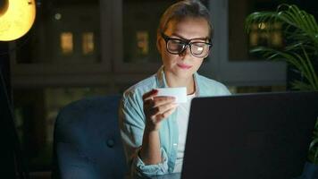 Woman is sitting in the armchair and makes an online purchase using a credit card and laptop at night video