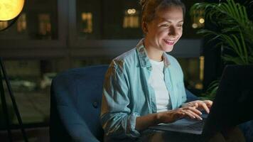 sorridente donna è seduta nel il poltrona e Lavorando su un' il computer portatile o chat con qualcuno a notte. concetto di a distanza opera video