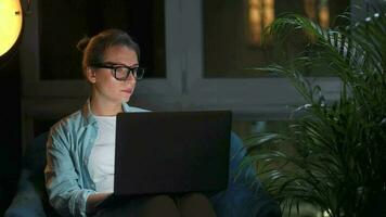 Frau mit Brille ist Sitzung im das Sessel und Arbeiten auf ein Laptop beim Nacht. Konzept von Fernbedienung Arbeit video