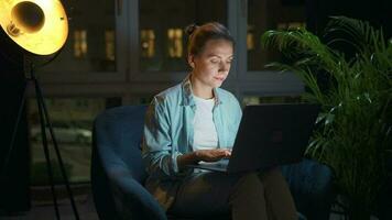 Woman is sitting in the armchair and working on a laptop at night. Concept of remote work video