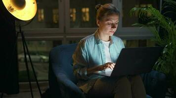 donna è seduta nel il poltrona e Lavorando su un' il computer portatile a notte. concetto di a distanza opera video