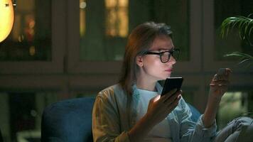 Woman is sitting in the armchair and makes an online purchase using a credit card and smartphone at night video