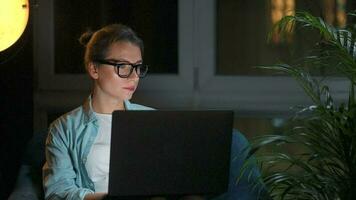 mujer con lentes es sentado en el Sillón y trabajando en un ordenador portátil a noche. concepto de remoto trabajo video